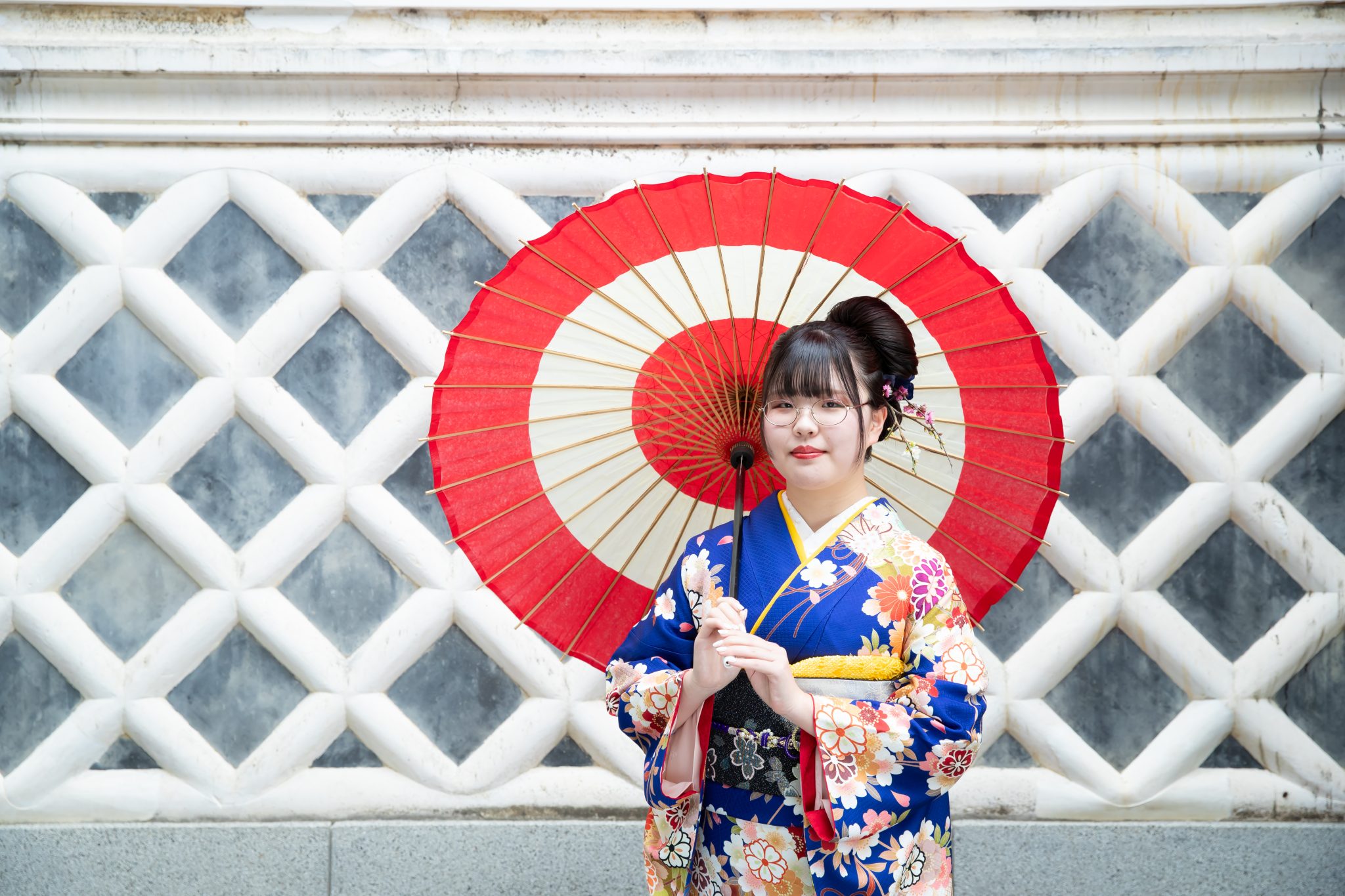 なまこ壁の前で和傘をさす青い振袖を着た女性